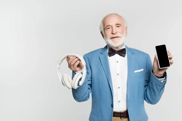 Hombre mayor sonriente sosteniendo auriculares y teléfonos inteligentes aislados en gris - foto de stock