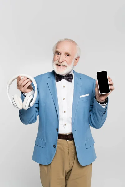 Позитивний старший чоловік у краватці та піджаку тримає навушники та смартфон із порожнім екраном ізольовано на сірому — стокове фото
