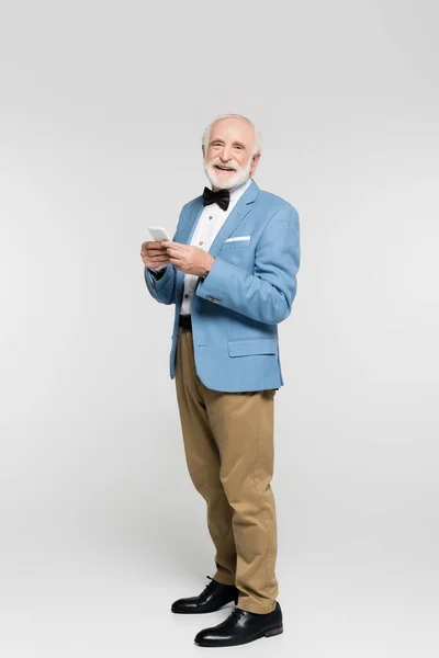 Hombre viejo sonriente en corbata de lazo y chaqueta que sostiene el teléfono inteligente sobre fondo gris - foto de stock