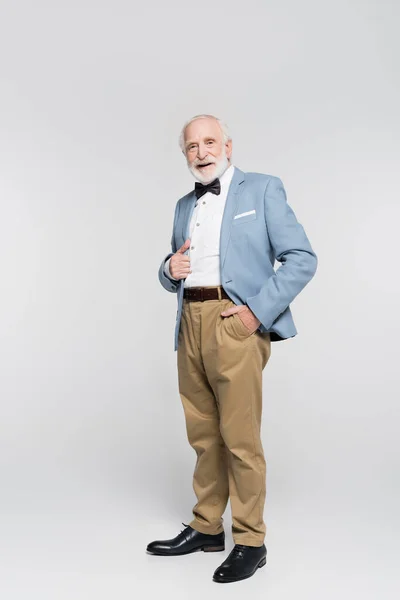 Hombre mayor sonriente en corbata de lazo y chaqueta azul sosteniendo la mano en el bolsillo de los pantalones sobre fondo gris - foto de stock