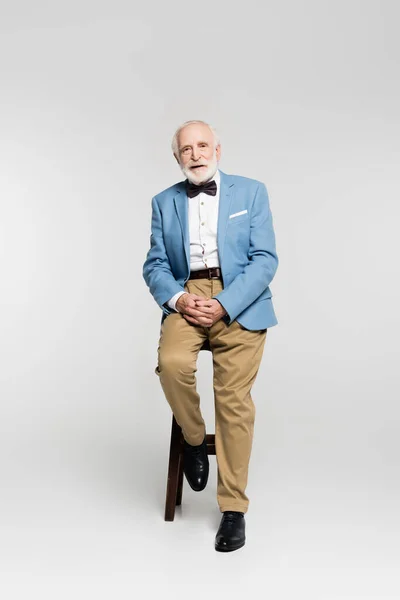 Homme âgé en nœud papillon et veste bleue assis sur une chaise sur fond gris — Photo de stock