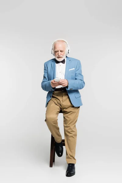 Senior man using headphones and smartphone on chair on grey background — Stock Photo