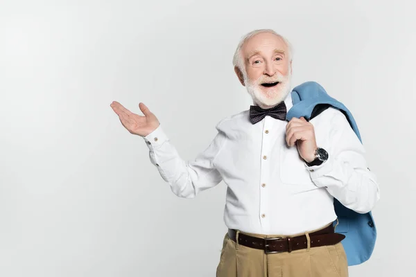 Homme âgé souriant en nœud papillon tenant veste et pointant à la main isolé sur gris — Photo de stock