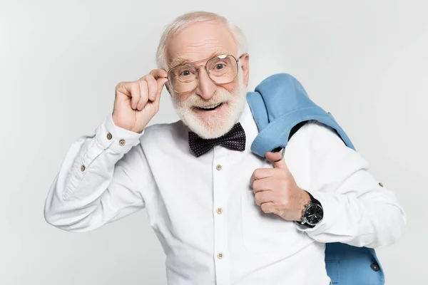Positive senior man in bow tie holding eyeglasses and jacket isolated on grey — Stock Photo
