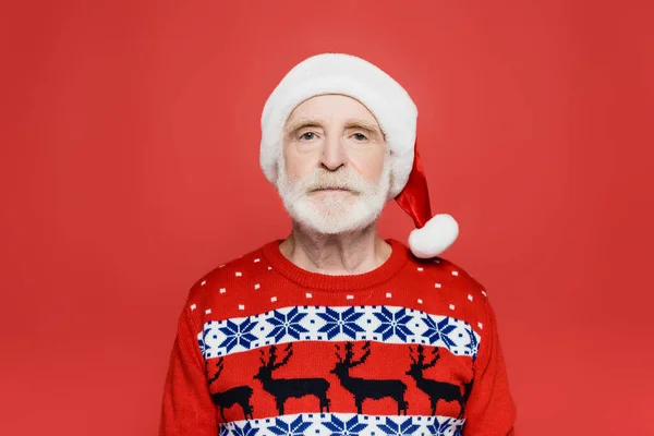 Homme âgé en bonnet santa et pull tricoté regardant la caméra isolée sur rouge — Photo de stock