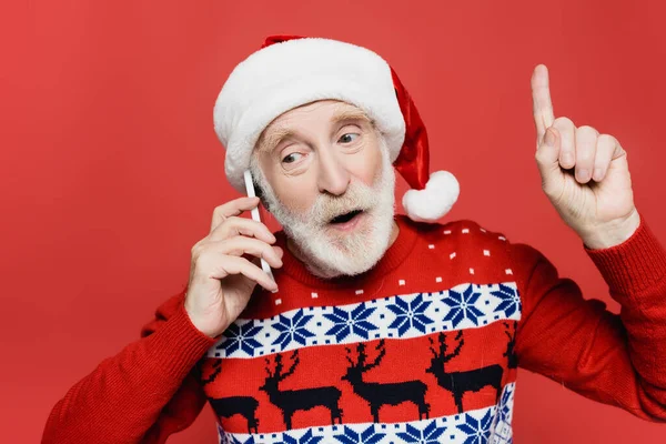 Senior man in santa hat talking on smartphone and pointing with finger isolated on red — Stock Photo