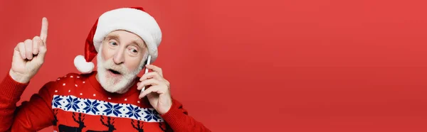 Elderly man in santa hat talking on smartphone and pointing with finger isolated on red, banner — Stock Photo