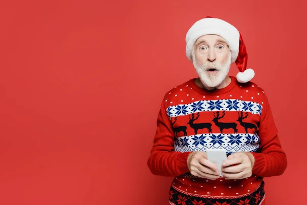 Shocked senior man in santa hat holding smartphone isolated on red — Stock Photo