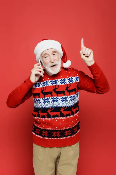 Hombre mayor en jersey de punto y sombrero de santa señalando con el dedo mientras habla en el teléfono inteligente sobre fondo rojo - foto de stock