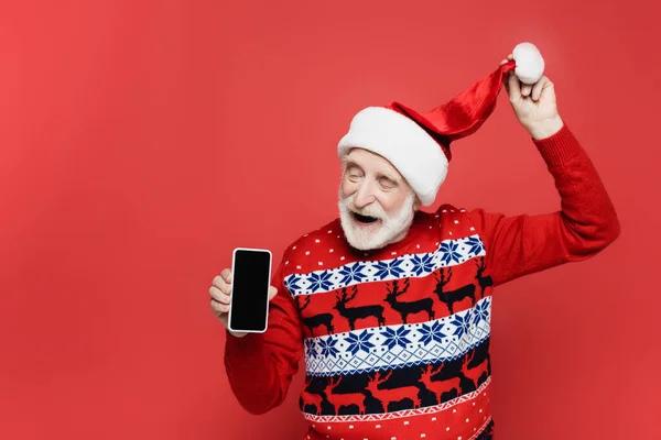 Positiver Senior zeigt Smartphone mit leerem Bildschirm und hält Weihnachtsmütze auf rotem Hintergrund — Stockfoto