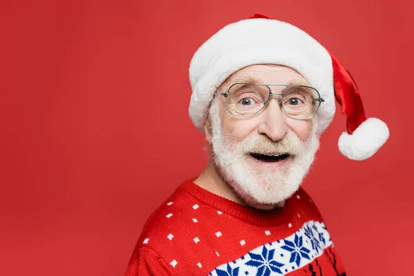 Hombre mayor en gafas y sombrero de santa sonriendo a la cámara aislado en rojo - foto de stock