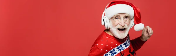 Allegro uomo anziano in cappello da Babbo Natale e cuffie che ballano isolate sul rosso, striscione — Foto stock