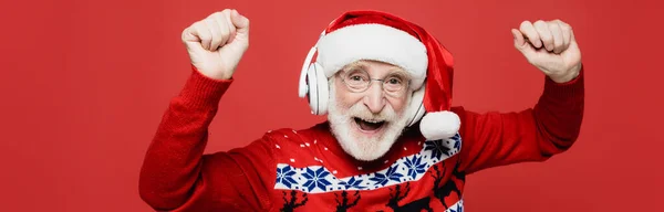 Allegro uomo anziano in cappello da Babbo Natale che balla ascoltando musica in cuffie isolate su rosso, striscione — Foto stock