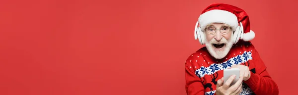 Allegro uomo anziano in cappello di Babbo Natale e cuffie utilizzando smartphone isolato su rosso, banner — Foto stock