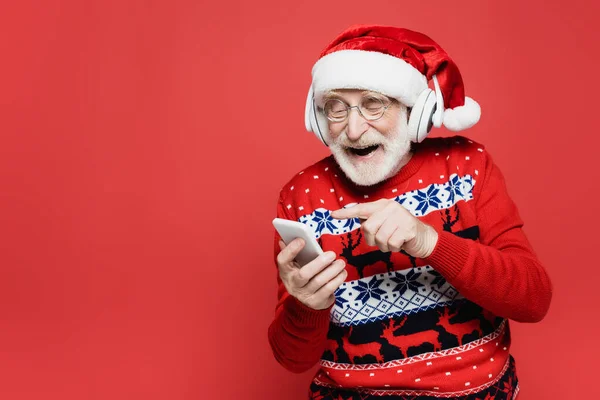 Lächelnder älterer Mann mit Kopfhörer und Weihnachtsmütze, der mit dem Finger auf ein rotes Smartphone zeigt — Stockfoto