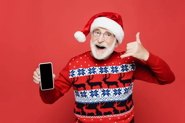 Hombre mayor alegre en sombrero de santa y suéter que muestra el teléfono inteligente con pantalla en blanco y gesto aislado en rojo - foto de stock
