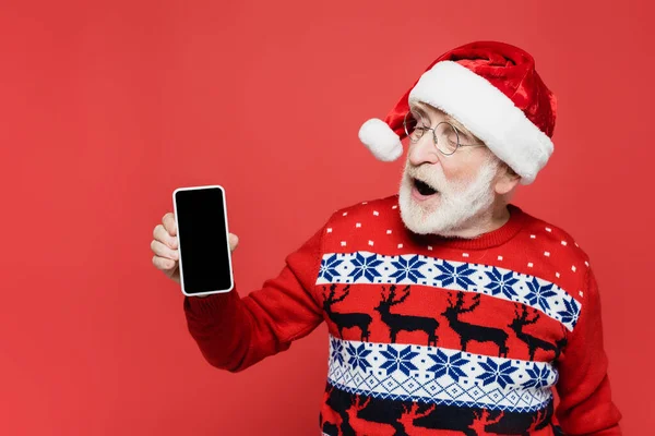 Захоплений старший чоловік у капелюсі Санта тримає смартфон з порожнім екраном ізольовано на червоному — стокове фото