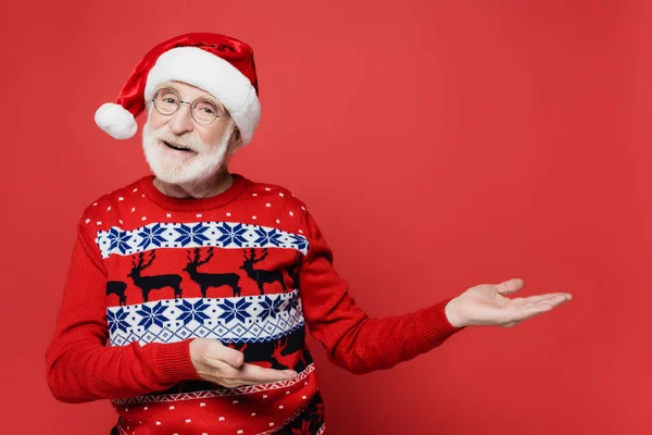 Smiling elderly man in sweater and santa hat pointing with hands on red background — Stock Photo