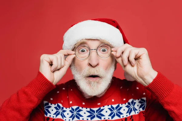Shocked senior man in santa hat and eyeglasses looking at camera isolated on red — Stock Photo