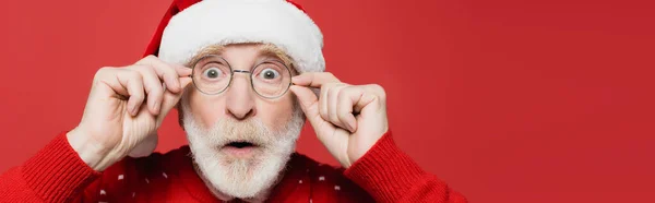 Surprised senior man in santa hat and eyeglasses looking at camera isolated on red, banner — Stock Photo