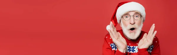 Sconvolto uomo anziano in cappello di Babbo Natale e maglione guardando la fotocamera isolata sul rosso, banner — Foto stock