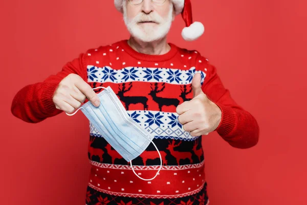 Vista ritagliata di uomo anziano in cappello di Babbo Natale mostrando pollice in su e tenendo maschera medica su sfondo sfocato isolato su rosso — Foto stock