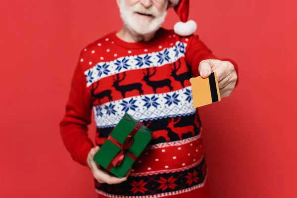 Cropped view of credit card in hand of senior man in santa hat with present on blurred foreground on red background — Stock Photo