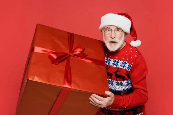 Homme âgé excité en santa chapeau regardant la caméra tout en tenant présent isolé sur rouge — Photo de stock