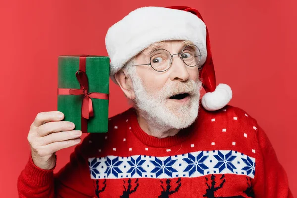 Excited elderly man in eyeglasses and santa hat holding present isolated on red — Stock Photo