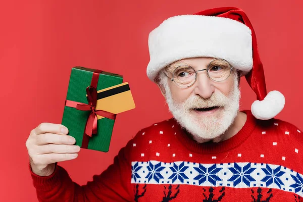 Літній чоловік в капелюсі Санта і в'язаний светр тримає подарункову коробку і кредитну картку ізольовано на червоному — стокове фото