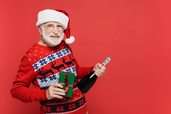 Uomo anziano in cappello da Babbo Natale sorridente alla macchina fotografica mentre tiene in mano una bottiglia di champagne e presente isolato sul rosso — Foto stock