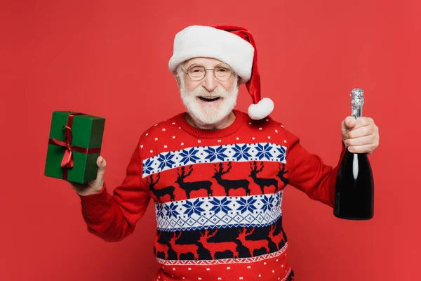 Positive senior man in santa hat and sweater holding bottle of champagne and gift on red background — Stock Photo