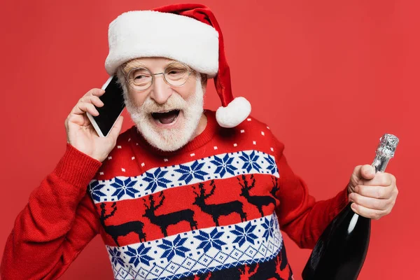 Joyeux homme âgé en chapeau de Père Noël parlant sur smartphone tout en tenant une bouteille de champagne isolé sur rouge — Photo de stock