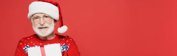 Uomo anziano sorridente in cappello di Babbo Natale tenuta presente isolato sul rosso, striscione — Foto stock