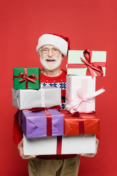 Positive elderly man in santa hat holding gift boxes isolated on red — Stock Photo