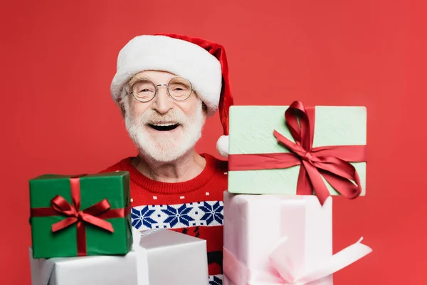 Alegre hombre mayor en sombrero de santa mirando a la cámara cerca de regalos en primer plano borroso aislado en rojo - foto de stock