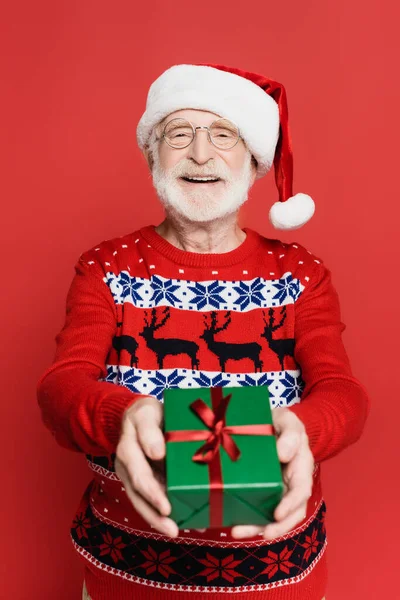 Smiling senior man in santa hat and knitted sweater showing gift box isolated on red — Stock Photo