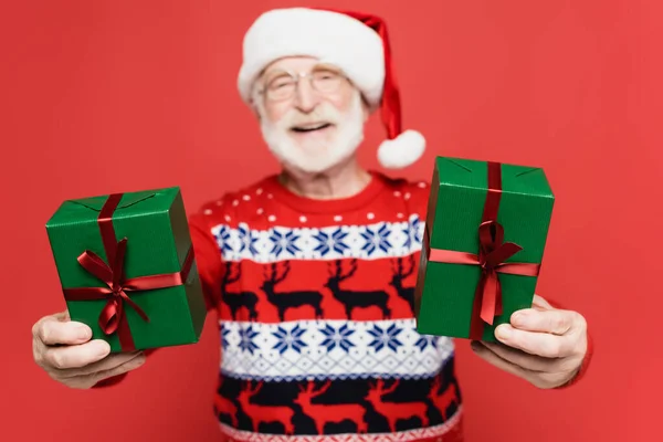 Gift boxes in hands of smiling senior man in santa hat on blurred background isolated on red — Stock Photo