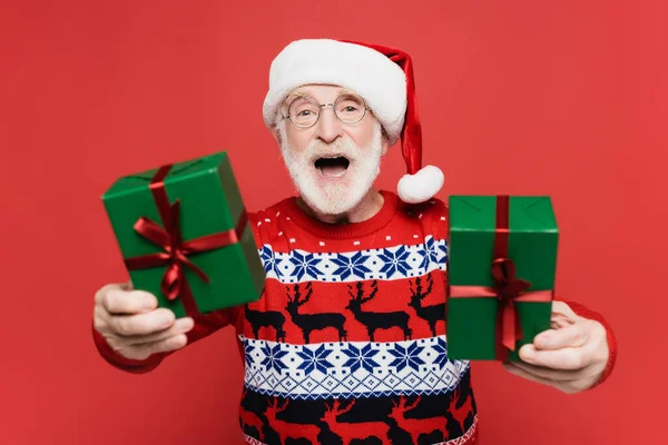 Alegre anciano en santa hat sosteniendo cajas de regalo en primer plano borroso aislado en rojo - foto de stock