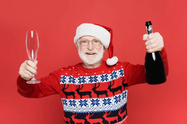 Homme âgé souriant en santa chapeau tenant bouteille de champagne et verre isolé sur rouge — Photo de stock