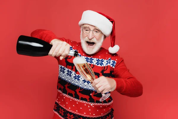 Vieil homme excité dans un chapeau de Père Noël versant du champagne dans un verre isolé sur rouge — Photo de stock