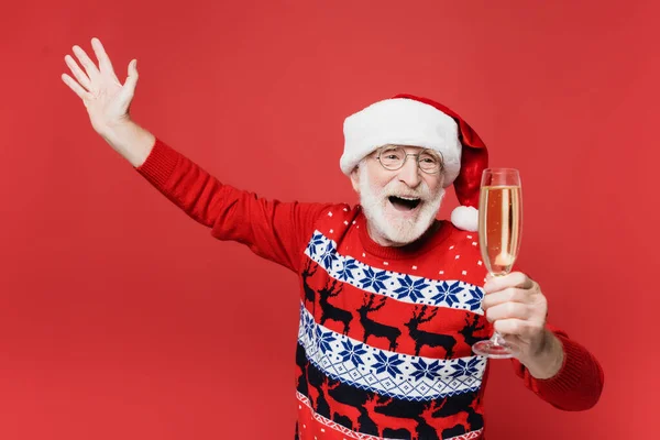 Homme âgé excité dans un chapeau de Père Noël tenant un verre de champagne isolé sur rouge — Photo de stock