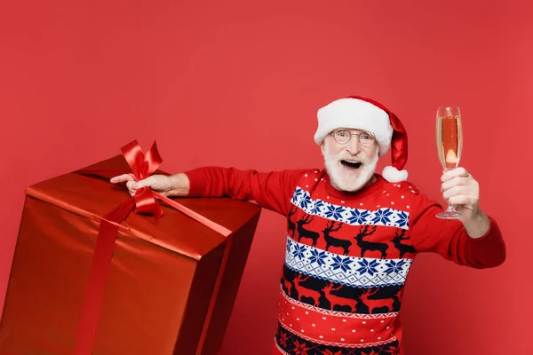 Cheerful senior man in santa hat holding glass of champagne and gift on red background — Stock Photo