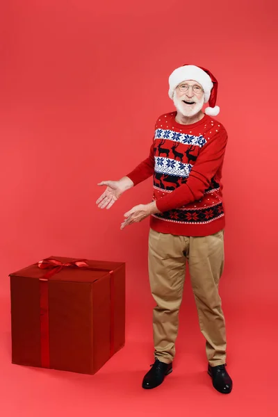 Smiling senior man in santa hat pointing at presents on red background — Stock Photo