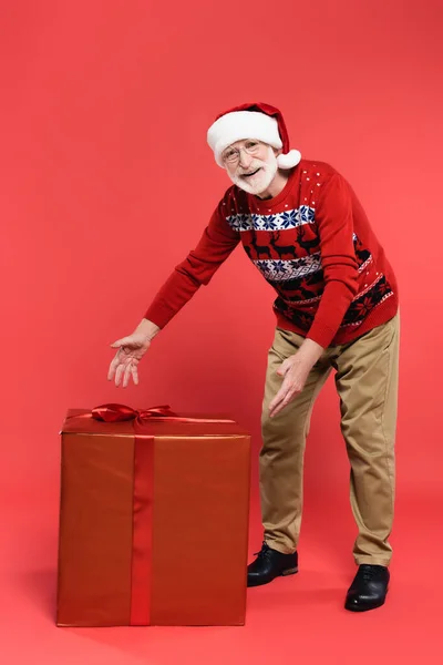 Cheerful senior man in santa hat and knitted sweater pointing at gift box on red background — Stock Photo