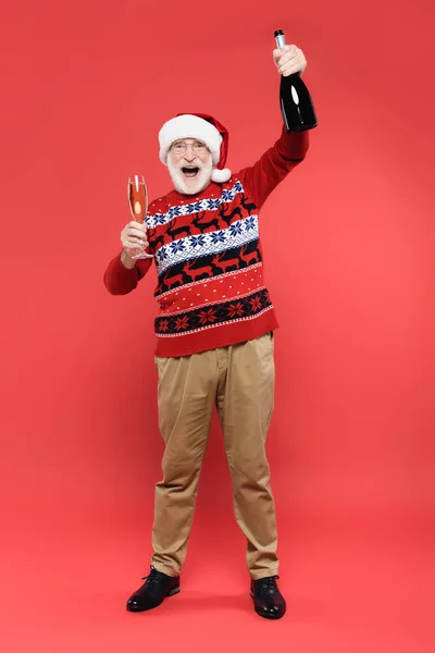 Positivo uomo anziano in cappello di Babbo Natale in possesso di vetro e bottiglia di champagne su sfondo rosso — Foto stock