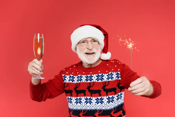 Homme âgé souriant au chapeau de Père Noël tenant un verre de champagne et scintillant isolé sur rouge — Photo de stock