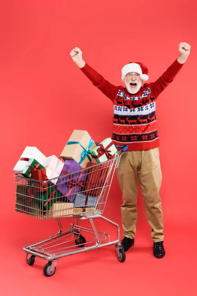 Aufgeregte Seniorin mit Weihnachtsmütze zeigt Yah-Geste in der Nähe des Einkaufswagens mit Geschenken auf rotem Hintergrund — Stockfoto