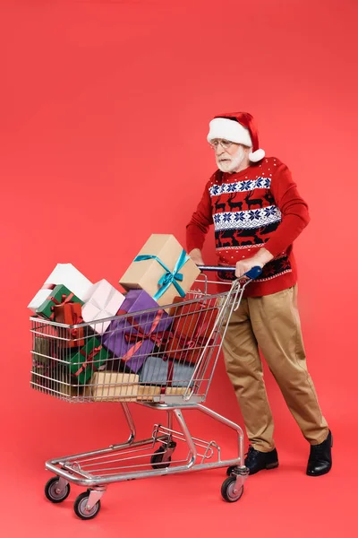 Homme âgé en pull et chapeau de Père Noël debout près du panier avec des cadeaux sur fond rouge — Photo de stock