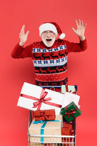 Shocked elderly man in santa hat looking at camera near presents in shopping cart isolated on red — Stock Photo
